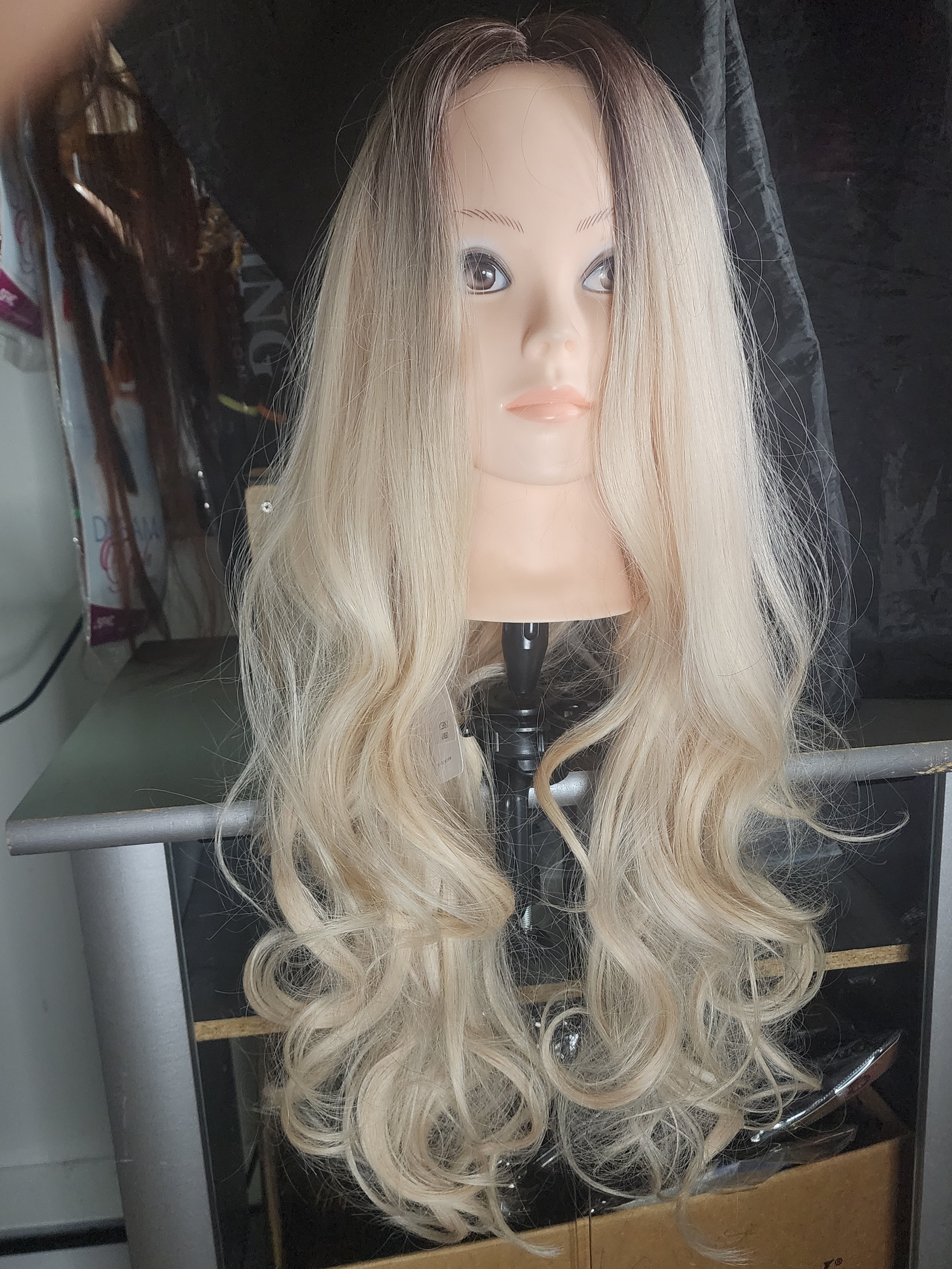 Long wavy wig with a blonde ombre color, displayed on a mannequin head stand in what appears to be a salon or studio setting.